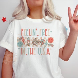 Op de afbeelding: Wit T-shirt met een rood, wit en blauwe afbeelding met de tekst "Feelin' Free * in the U.S.A. *" met sterren en andere patriottische afbeeldingen.