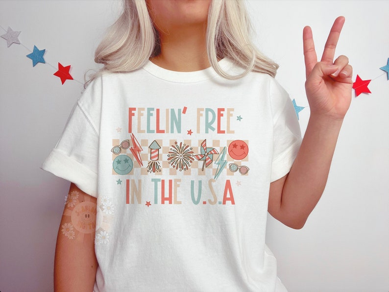 Op de afbeelding: Wit T-shirt met een rood, wit en blauwe afbeelding met de tekst "Feelin' Free * in the U.S.A. *" met sterren en andere patriottische afbeeldingen.
