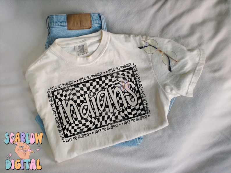 May include: A cream-coloured t-shirt featuring the word "Indians" in a black and white chequered design. The shirt is folded over blue jeans, with round-framed glasses on top. The text "lets go indians" is printed around the design.