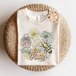 May include: A beige t-shirt with a graphic of a bee and flowers in a honeycomb pattern. The text "Sweet Like Honey" is printed in a retro font.