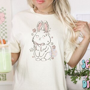 May include: A white t-shirt with a cute cartoon bunny wearing a pink bow and surrounded by pink flowers. The bunny is sitting down and has a happy expression on its face.