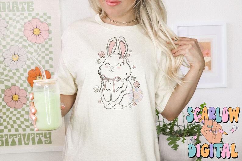 May include: A white t-shirt with a cute cartoon bunny wearing a pink bow and surrounded by pink flowers. The bunny is sitting down and has a happy expression on its face.