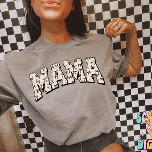 Puede incluir: Una camiseta gris con la palabra "MAMA" impresa en blanco y negro. Las letras est&aacute;n formadas por un patr&oacute;n de peque&ntilde;os lazos negros y blancos.