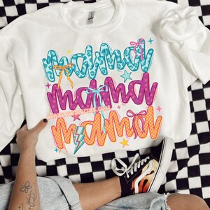 May include: White sweatshirt with the text "Mama Mama Mama" in colorful, stylized lettering. The text is decorated with stars, lightning bolts, and bows.