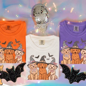 K&ouml;nnte beinhalten: Drei wei&szlig;e T-Shirts mit einem Halloween-Motiv, das drei Golden Retriever zeigt, die Hexenh&uuml;te und Schleifen tragen. Die Shirts werden auf einem lila und rosa Hintergrund mit einer Discokugel pr&auml;sentiert.