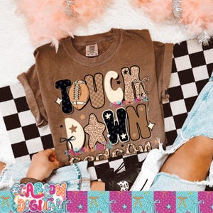 May include: A brown t-shirt with the words "TOUCH DOWN" in a mix of fonts and patterns, including a football graphic and star shapes. The shirt is a casual style, likely for everyday wear.