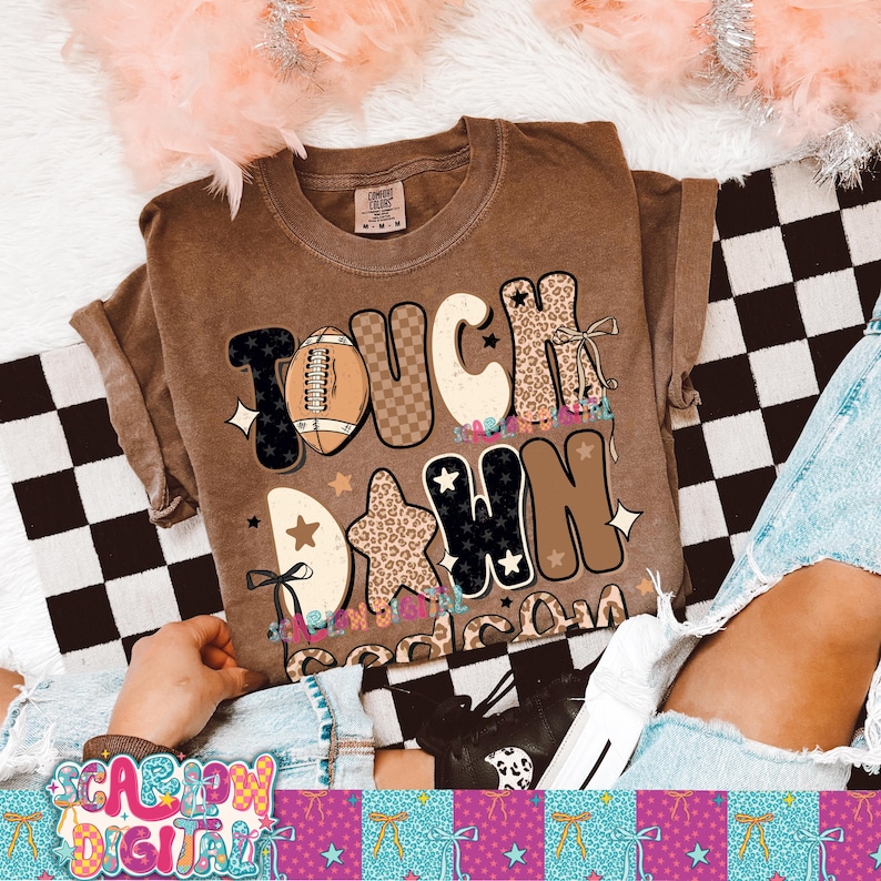May include: A brown t-shirt with the words "TOUCH DOWN" in a mix of fonts and patterns, including a football graphic and star shapes. The shirt is a casual style, likely for everyday wear.