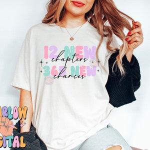 May include: A white t-shirt with the text "12 new chapters + 365 new chances" in colorful, bubbly letters. The text is decorated with stars and a small floral design.