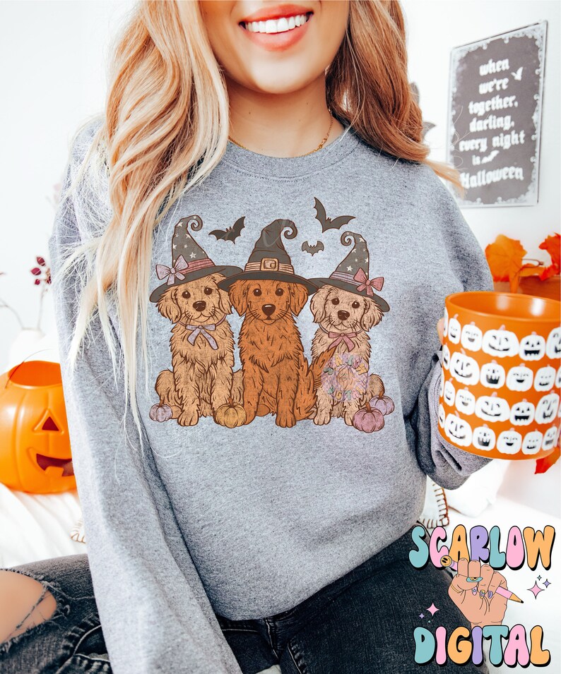 K&ouml;nnte beinhalten: Ein graues Sweatshirt mit einem Motiv von drei Golden Retrievern, die Hexenh&uuml;te tragen und K&uuml;rbisse halten. Die Hunde sitzen in einer Reihe und haben rosa Schleifen an ihren H&uuml;ten. Das Sweatshirt hat den Text "SCARLOW DIGITAL" in der rechten unteren Ecke.