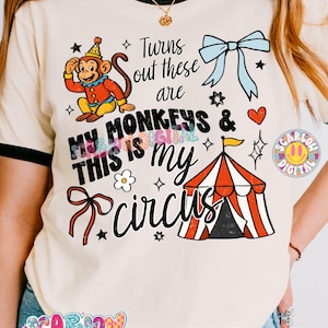 May include: Cream-colored t-shirt with black trim featuring a circus-themed graphic. The design includes a cartoon monkey, a circus tent, and the text "Turns out these are my monkeys & this is my circus."