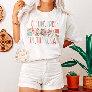 Op de afbeelding: Wit T-shirt met de tekst "Feelin' Free* in the U.S.A." in roze, blauw en rood. De tekst wordt omringd door een geruit patroon en afbeeldingen van vuurwerk, zonnebrillen en een windmolen.