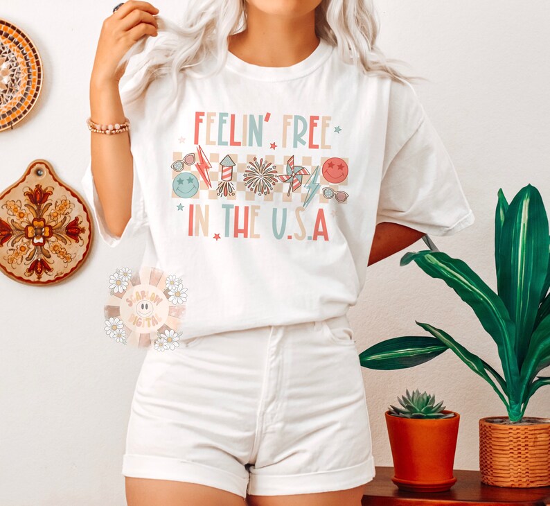 Op de afbeelding: Wit T-shirt met de tekst "Feelin' Free* in the U.S.A." in roze, blauw en rood. De tekst wordt omringd door een geruit patroon en afbeeldingen van vuurwerk, zonnebrillen en een windmolen.