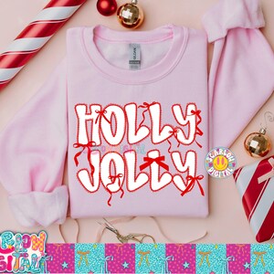 以下が含まれることがあります： 「HOLLY JOLLY」の文字が赤の縁取りとリボンで装飾された白いバブルレターで描かれた、明るいピンク色のスウェットシャツ。オーナメントや包装されたギフトなど、クリスマスをテーマにした装飾がスウェットシャツを囲んでいます。