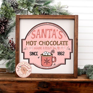 Op de afbeelding: Een roze en wit bord met de tekst "Santa's Hot Chocolate - Made Fresh Daily - Since 1862" en een rode mok met marshmallows en een snoepstok.