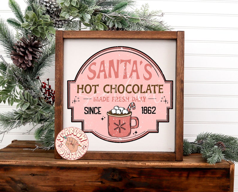Op de afbeelding: Een roze en wit bord met de tekst "Santa's Hot Chocolate - Made Fresh Daily - Since 1862" en een rode mok met marshmallows en een snoepstok.