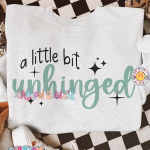 K&ouml;nnte beinhalten: Ein hellgraues Sweatshirt mit dem Schriftzug "a little bit unhinged" in Schwarz und T&uuml;rkis. Das Design enth&auml;lt Sternakzente. Das Sweatshirt wird auf einer karierten Oberfl&auml;che pr&auml;sentiert.