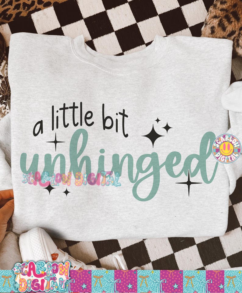 K&ouml;nnte beinhalten: Ein hellgraues Sweatshirt mit dem Schriftzug "a little bit unhinged" in Schwarz und T&uuml;rkis. Das Design enth&auml;lt Sternakzente. Das Sweatshirt wird auf einer karierten Oberfl&auml;che pr&auml;sentiert.