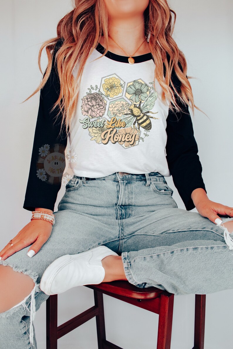 May include: A white and black baseball tee shirt with a graphic of a honeycomb with flowers and a bee. The text "Sweet Like Honey" is printed on the shirt. The model is wearing blue jeans and white sneakers.