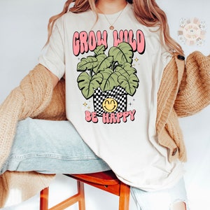 May include: A white t-shirt with a pink and green graphic that says "Grow Wild Be Happy". The graphic features a potted plant with a smiley face.