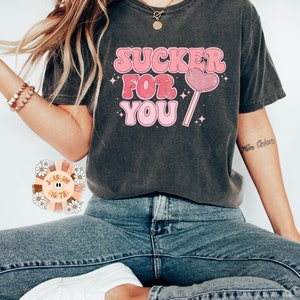 Może przedstawiać: Szary t-shirt z nadrukiem w kolorze r&oacute;żowym i białym, na kt&oacute;rym widnieje napis "SUCKER FOR YOU" oraz r&oacute;żowy lizak w kształcie serca.