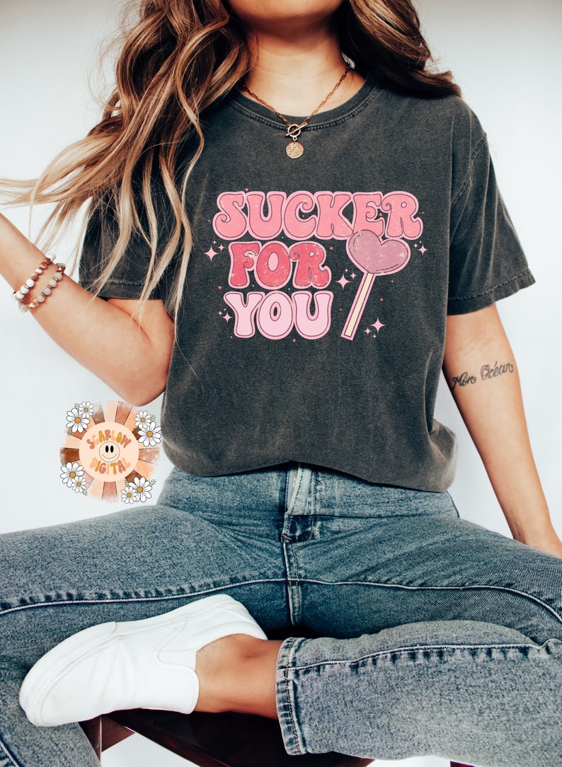 Może przedstawiać: Szary t-shirt z nadrukiem w kolorze r&oacute;żowym i białym, na kt&oacute;rym widnieje napis "SUCKER FOR YOU" oraz r&oacute;żowy lizak w kształcie serca.