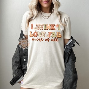 Op de afbeelding: Een beige T-shirt met de tekst "I think I love fall most of all" in kleurrijke, gepatroneerde letters.