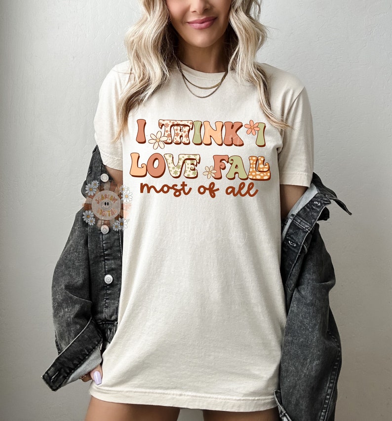 Op de afbeelding: Een beige T-shirt met de tekst "I think I love fall most of all" in kleurrijke, gepatroneerde letters.