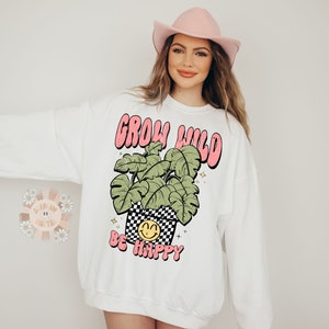 May include: White sweatshirt with a pink and green graphic of a plant in a pot with a smiley face and the text "Grow Wild Be Happy".