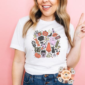 Peut inclure: Un t-shirt blanc avec un motif color&eacute; sur le th&egrave;me d'Halloween repr&eacute;sentant des fant&ocirc;mes, des citrouilles, des chauves-souris, des bonbons et le texte "Boo!" et "Trick or Treat".