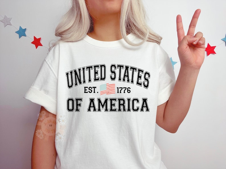 May include: White t-shirt with black text that reads "United States Est. 1776 Of America". A small American flag is printed next to the year.