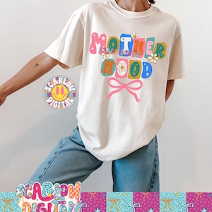 Puede incluir: Camiseta color crema con la palabra "MOTHERHOOD" en letras coloridas de estilo retro. El dise&ntilde;o incluye flores y un lazo rosa. La camiseta se combina con unos vaqueros azules.