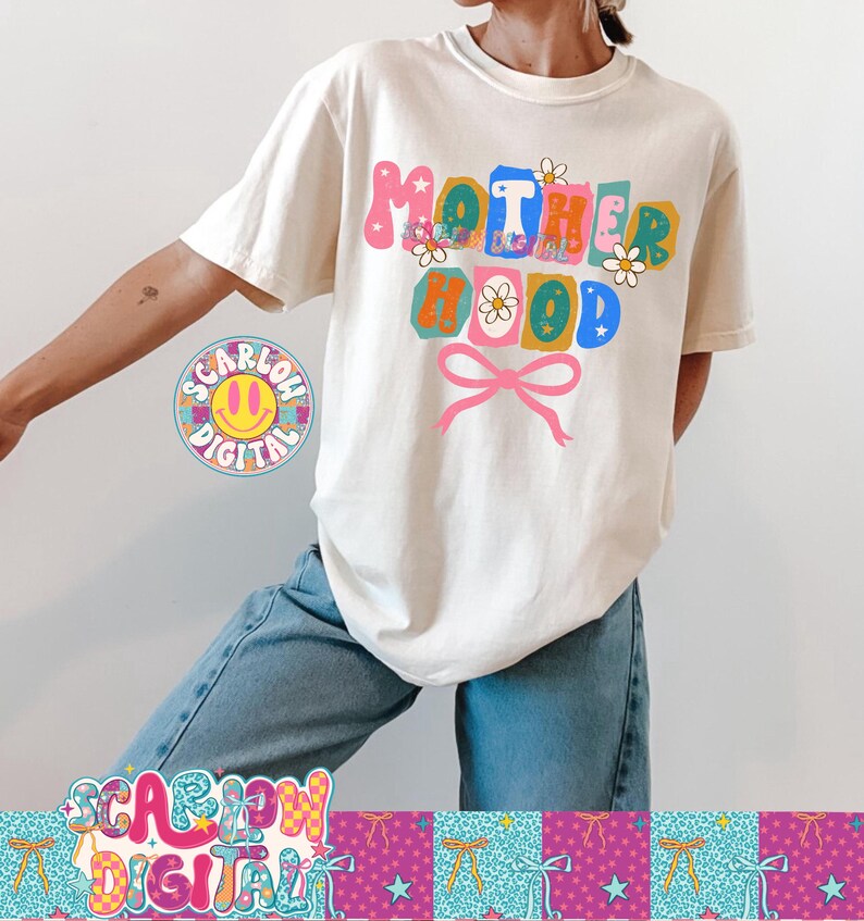 Puede incluir: Camiseta color crema con la palabra "MOTHERHOOD" en letras coloridas de estilo retro. El dise&ntilde;o incluye flores y un lazo rosa. La camiseta se combina con unos vaqueros azules.