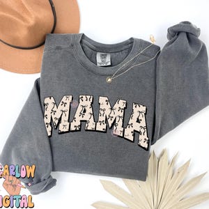 Puede incluir: Una sudadera gris con la palabra "MAMA" impresa en letras grandes y en negrita. Las letras est&aacute;n rellenas con un patr&oacute;n de lazos rosas con detalles negros.