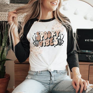 May include: A white and black baseball tee shirt with the text "Spooky Vibes" in a retro font. The text is surrounded by skeleton hands and flowers. The shirt is tied at the waist.
