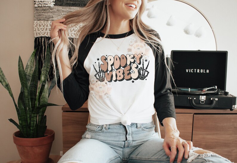 May include: A white and black baseball tee shirt with the text "Spooky Vibes" in a retro font. The text is surrounded by skeleton hands and flowers. The shirt is tied at the waist.
