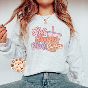 May include: A light gray sweatshirt with the text "Half Human Half Coffee" in a retro font, accompanied by a graphic of an iced coffee. The design features pink, orange, and brown tones. The sweatshirt is paired with blue jeans.