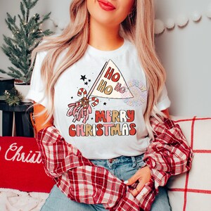 May include: White t-shirt with a red, pink, and gold graphic design. The design features a pom-pom, candy canes, and a flag that says "Ho Ho Ho" and "Merry Christmas".