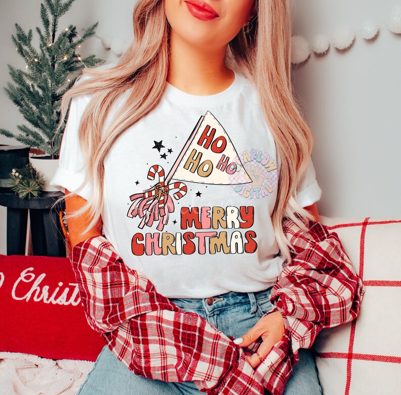 May include: White t-shirt with a red, pink, and gold graphic design. The design features a pom-pom, candy canes, and a flag that says "Ho Ho Ho" and "Merry Christmas".