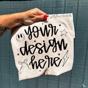 Könnte beinhalten: Ein hellfarbenes T-Shirt mit dem Schriftzug "YOUR DESIGN HERE" in schwarzer Schrift. Das Design umfasst ein Smiley, Sterne, einen Stern, eine Schleife und einen Blitz. Das T-Shirt wird von einer Hand gehalten.
