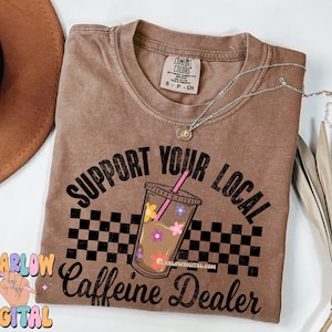 May include: A brown t-shirt with the text "Support Your Local Caffeine Dealer" in black. The text is surrounded by a checkered pattern and a cartoon image of a cup of iced coffee with flowers.