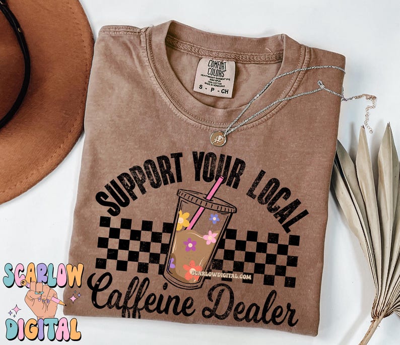 May include: A brown t-shirt with the text "Support Your Local Caffeine Dealer" in black. The text is surrounded by a checkered pattern and a cartoon image of a cup of iced coffee with flowers.