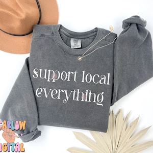 May include: A dark gray sweatshirt with the text "support local everything" printed in white on the front. The sweatshirt is made of a soft, comfortable material and has a relaxed fit.