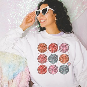 May include: White sweatshirt with a colorful design of nine disco balls. The disco balls are arranged in a 3x3 grid pattern. The sweatshirt has a crew neck and long sleeves.