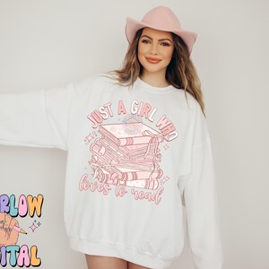 Puede incluir: Sudadera blanca con el texto "Just a girl who loves to read" y una pila de libros rosas con un dise&ntilde;o floral.