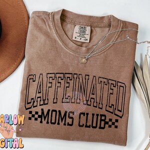 K&ouml;nnte beinhalten: Ein braunes T-Shirt mit dem Text "CAFFEINATED MOMS CLUB" in schwarzen Buchstaben. Das Shirt hat einen Used-Look und ist aus einem weichen, bequemen Stoff.