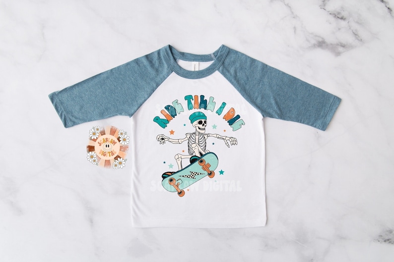 K&ouml;nnte beinhalten: Wei&szlig;es und blaues Baseball-T-Shirt mit einem Skelett, das Skateboard f&auml;hrt, und dem Text "Ride Til I Die".