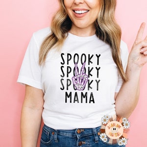 K&ouml;nnte beinhalten: Wei&szlig;es T-Shirt mit schwarzem Text, der "Spooky Spooky Spooky Mama" lautet, und einer lila Skeletthand, die ein Peace-Zeichen macht.