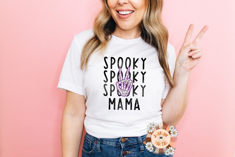 K&ouml;nnte beinhalten: Wei&szlig;es T-Shirt mit schwarzem Text, der "Spooky Spooky Spooky Mama" lautet, und einer lila Skeletthand, die ein Peace-Zeichen macht.