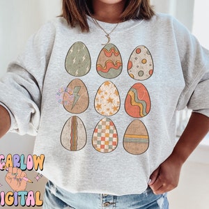 May include: A gray sweatshirt with a pattern of nine colorful Easter eggs. Each egg has a different design, including stripes, polka dots, stars, and lightning bolts.