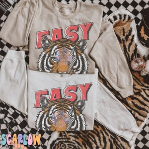 Puede incluir: Dos camisas beige de manga larga con un gr&aacute;fico de tigre y la palabra "EASY" en letras rojas. El tigre tiene un parche en el ojo a cuadros y detalles coloridos.